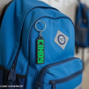 Könnte beinhalten: Ein blauer Rucksack mit einem schwarz-grünen Schlüsselanhänger mit der Aufschrift "CINDY" in pixeligen grünen Buchstaben. Der Schlüsselanhänger ist am Reißverschluss des Rucksacks befestigt. Ein weiterer Rucksack ist im Hintergrund zu sehen.