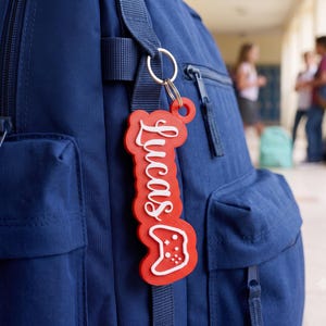 Könnte beinhalten: Ein roter Acryl-Schlüsselanhänger an einem marineblauen Rucksack. Der Anhänger zeigt den Namen Lucas in weißer Schreibschrift über einem weißen Umriss eines Videospiel-Controllers. Das Design ist mehrschichtig mit einer roten Basis.