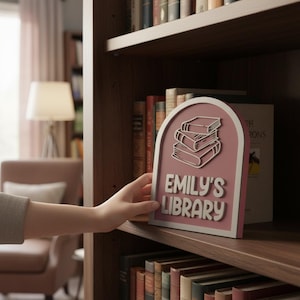 May include: A pink and white sign with the text "EMILY'S LIBRARY" and a stack of books graphic. The sign is being placed on a wooden bookshelf filled with books. The sign has a rounded top.
