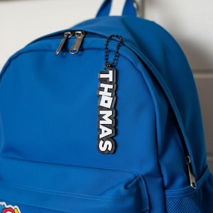 Puede incluir: Una mochila azul con una etiqueta con el nombre "THOMAS" en blanco y negro. La etiqueta está unida a la mochila con una cadena. La mochila tiene un compartimento principal con cremallera y un bolsillo frontal.