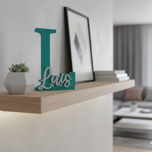 Könnte beinhalten: Ein türkisfarbener Holzbuchstabe "L" mit dem Namen "Luis" in weißer Schrift, der auf einem hellen Holzregal steht. Eine kleine Sukkulente in einem weißen geometrischen Topf und ein gerahmtes abstraktes Kunstwerk befinden sich ebenfalls auf dem Regal. Die Szene spielt in einem modernen Wohnraum.