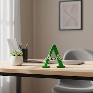 May include: A green wooden letter "A" with the name "Amelia" in cursive script. The letter is on a wooden desk, with a laptop, a small succulent plant in a white pot, and a framed picture in the background.