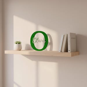 Op de afbeelding: Een groene houten letter "O" met de naam "Oliver" uitgesneden, weergegeven op een lichtgekleurde houten plank. De plank bevat ook een kleine potplant en verschillende boeken. De achtergrond is een neutrale muur.