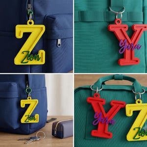 Puede incluir: Un conjunto de etiquetas personalizadas para bolsos con una letra Z amarilla con el nombre Zion en verde y una letra Y roja con el nombre Yara en morado. Estos dijes de plástico con letras están sujetos a mochilas con anillos de metal.