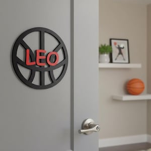 May include: A black basketball-themed wall decoration with the name "LEO" in red letters. The basketball design is mounted on a gray door. A basketball and framed art are visible in the background.