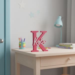 May include: A pink wooden letter "K" with the name "Kyla" in white script. The letter is on a light wood desk, with a blue desk lamp and a stack of books in the background. The room has a light blue door.