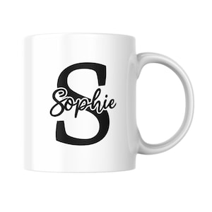May include: White ceramic mug with a large black letter "S" and the name "Sophie" in a cursive font. The mug has a curved handle and is designed for hot beverages.