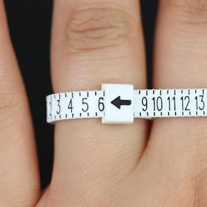 Può includere: Un misuratore di misura del dito in plastica bianca con numeri neri e una freccia. I numeri sul misuratore vanno da 2 a 15.