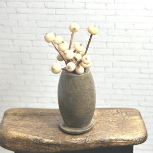 May include: A small, rustic wooden stool holds a weathered, gray-brown vase filled with cream-colored berry stems. The vase has a rounded base and a narrow neck. The background is a white brick wall.