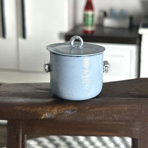 May include: A miniature light blue cooking pot with a lid and silver handles. The pot is sitting on a small, dark wooden stool. The background includes a miniature kitchen scene with a stove and cabinets.