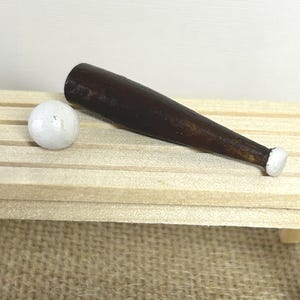May include: A miniature dark brown wooden baseball bat with a white painted knob and a small, round, white baseball. The bat has a smooth, polished finish and a tapered shape, while the baseball is spherical with a matte white surface texture.