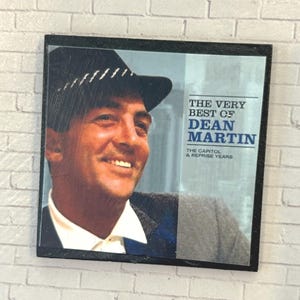 May include: A square album cover featuring a portrait of Dean Martin wearing a dark hat and suit. The text reads THE VERY BEST OF DEAN MARTIN THE CAPITOL & REPRISE YEARS. The image is framed in black and has a glossy finish with visible surface scratches.