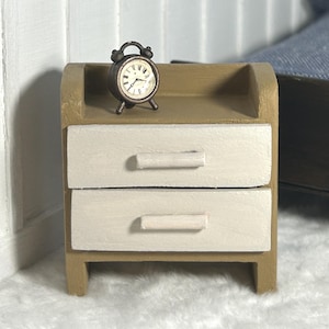 May include: Miniature wooden nightstand with two drawers painted cream and a light brown frame. A small, antique-style clock sits on top. The nightstand is part of a dollhouse bedroom set.