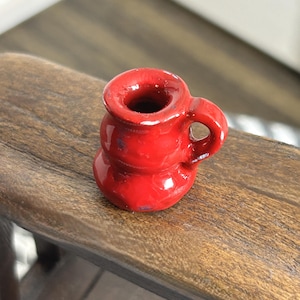 May include: A small, red ceramic mug with a handle. The mug has a rounded base and a flared rim. The exterior is a glossy red with some visible blue paint. The mug is sitting on a wooden surface.