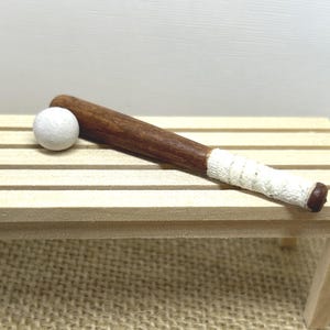 May include: A miniature wooden baseball bat with a dark brown finish and a white textured grip tape wrapped around the handle, positioned next to a small, round, solid white baseball. The items are crafted with a smooth, polished wood texture.