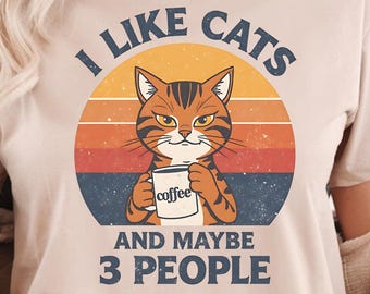 May include: Beige t-shirt with a retro design featuring a cat holding a coffee cup. The text reads "I LIKE CATS AND MAYBE 3 PEOPLE" in a vintage font, set against a sunset background. The cat is orange and brown.