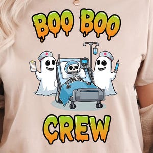 Può includere: T-shirt beige con grafica a tema Halloween. Il design presenta le parole "Boo Boo Crew" in verde e arancione, insieme a fantasmi dei cartoni animati e un paziente scheletrico su un letto d'ospedale. Un capo casual.