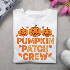 May include: White t-shirt with a pumpkin-themed design. The shirt features three orange jack-o'-lanterns above the words "PUMPKIN PATCH CREW" in orange and yellow lettering, with leaf accents. A festive fall design.