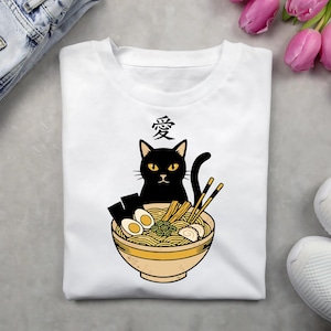 May include: White t-shirt featuring a black cat with yellow eyes, sitting in a bowl of ramen. The ramen bowl includes noodles, eggs, and chopsticks. Above the cat is the Japanese character for love.