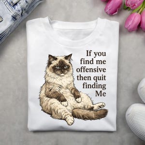 May include: White t-shirt featuring a detailed illustration of a fluffy cat with blue eyes. The text on the shirt reads, "If you find me offensive then quit finding Me."