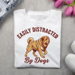 Op de afbeelding: Wit T-shirt met de tekst "EASILY DISTRACTED By Dogs" in een bruin, vintage lettertype. Een bruine en beige hondenillustratie staat eronder. Het shirt is opgevouwen op een grijze ondergrond met een spijkerbroek en bloemen.