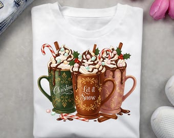 May include: White t-shirt featuring a festive design of three mugs of hot chocolate with whipped cream, chocolate drizzle, and candy canes. The mugs are green, brown, and pink, with the words "Christmas" and "Let it Snow".
