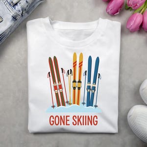 May include: White t-shirt with a graphic of three pairs of skis in red, yellow, and blue, with ski poles. The phrase "GONE SKIING" is printed in red below the skis. The shirt is on a gray surface.