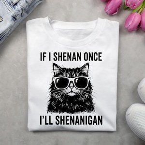May include: White t-shirt with a black graphic of a cat wearing sunglasses. The text above the cat reads "IF I SHENAN ONCE" and the text below reads "I'LL SHENANIGAN".