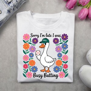 Puede incluir: Camiseta blanca con un ganso de dibujos animados que lleva un sombrero verde de "Entrenador" y sostiene una pelota de béisbol. El ganso está rodeado de flores coloridas y el texto "Lo siento, llego tarde, estaba ocupado bateando".