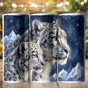 May include: Three stainless steel tumblers display a watercolour illustration of snow leopards against a dark blue starry night sky. The design includes geometric mountain peaks and snowflakes, creating a winter theme.