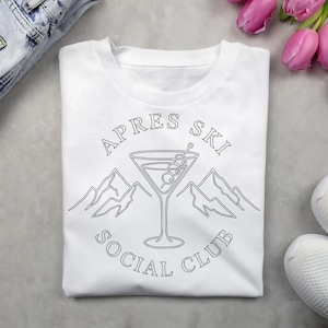 May include: White t-shirt with a black graphic design. The design features a martini glass with olives, mountains, and the text "APRES SKI SOCIAL CLUB" in a circular layout. The shirt is folded on a gray surface.