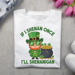 May include: White t-shirt featuring a cartoon leprechaun in a green hat and suit, green sunglasses, and a red beard. The shirt has the text "IF I SHENAN ONCE I'LL SHENANIGAN" and a pot of gold.