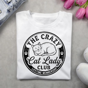 May include: White t-shirt featuring a black and white graphic. The graphic is a circular design with the words "THE CRAZY Cat Lady CLUB LIFETIME MEMBERSHIP" surrounding a sleeping cat illustration and paw prints.