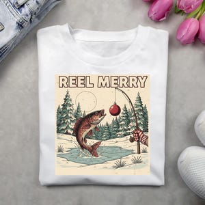 May include: White t-shirt with a graphic featuring a jumping fish, a fishing rod with a red ornament, and the text "REEL MERRY." The design includes a winter landscape with pine trees and a full moon.