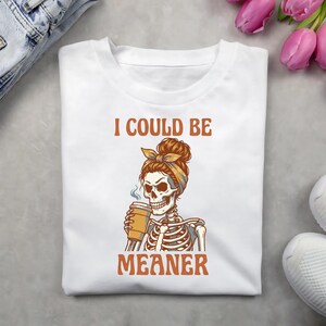 May include: White t-shirt with the text "I COULD BE MEANER" in orange. The graphic features a skeleton with a bun and bandana holding a coffee cup. The shirt is folded on a light gray surface.
