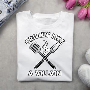 Può includere: T-shirt bianca con una grafica che dice "Grillin' Like A Villain". Il design presenta utensili da griglia incrociati: una spatola e una forchetta, con un sbuffo di fumo. Il testo e la grafica sono in nero.