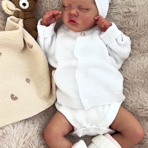 May include: A lifelike baby doll dressed in a white knitted cardigan, diaper, and booties. The doll wears a matching white hat with a pink bow. A brown crocheted teddy bear and a beige blanket with a rainbow design are nearby.