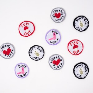 May include: A collection of embroidered patches with various designs and text. The patches are circular, with white, red, black, and gray borders. Some feature a heart, a mushroom, a bookworm, and a bug, with phrases like "Library Lover" and "Don't Bug Me!"
