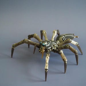 May include: A bronze and silver mechanical spider sculpture. The spider has multiple articulated legs and a segmented body with intricate detailing. The sculpture has blue accents and is set against a grey background.