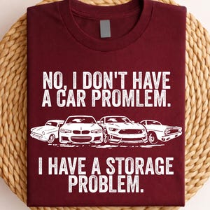 May include: Maroon t-shirt with white text and car graphics. The text reads "NO, I DON'T HAVE A CAR PROBLEM. I HAVE A STORAGE PROBLEM." The shirt is folded on a woven, circular mat.