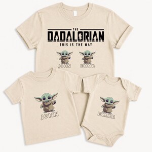 May include: Beige t-shirts and a babygrow featuring a green alien character. The adult shirt reads "The Dadalorian, This is the Way." The children's shirts have the names "John" and "Emma" below the character.