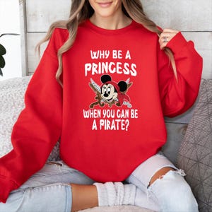 K&ouml;nnte beinhalten: Ein rotes Sweatshirt mit dem Text "WHY BE A PRINCESS WHEN YOU CAN BE A PIRATE?" und einer Cartoon-Grafik einer Piratenmaus. Die Grafik zeigt einen Piratenhut, eine Augenklappe und gekreuzte Schwerter.