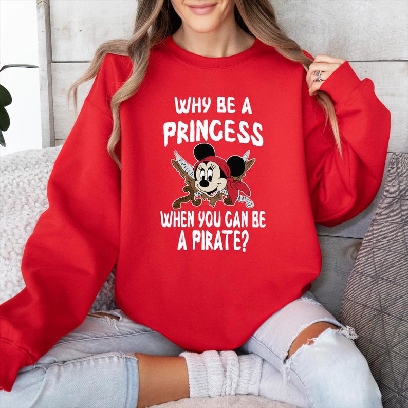 K&ouml;nnte beinhalten: Ein rotes Sweatshirt mit dem Text "WHY BE A PRINCESS WHEN YOU CAN BE A PIRATE?" und einer Cartoon-Grafik einer Piratenmaus. Die Grafik zeigt einen Piratenhut, eine Augenklappe und gekreuzte Schwerter.