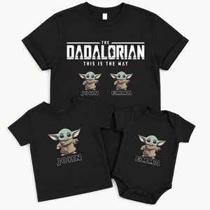 May include: Black t-shirts and a babygrow featuring the text "The Dadalorian This Is The Way" and cartoon images of a green alien. The shirts are personalised with the names "John" and "Emma".