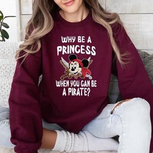K&ouml;nnte beinhalten: Burgunderfarbenes Sweatshirt mit dem Text "WHY BE A PRINCESS WHEN YOU CAN BE A PIRATE?" und einer Grafik einer Piratenmaus. Die Maus tr&auml;gt ein rotes Bandana und h&auml;lt gekreuzte Schwerter.