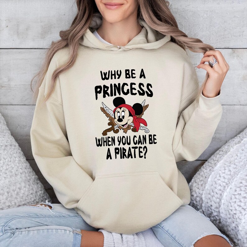 K&ouml;nnte beinhalten: Beigefarbener Kapuzenpullover mit dem Text "WHY BE A PRINCESS WHEN YOU CAN BE A PIRATE?" und einem Cartoon-Maus-Motiv mit rotem Bandana und gekreuzten Schwertern. Der Hoodie hat eine Vordertasche und eine Kapuze mit Kordelzug.