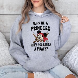 K&ouml;nnte beinhalten: Hellgrauer Kapuzenpullover mit dem Text "WHY BE A PRINCESS WHEN YOU CAN BE A PIRATE?". Das Design zeigt eine Cartoon-Maus mit Piratenhut und gekreuzten Schwertern. Ein l&auml;ssiger Stil.