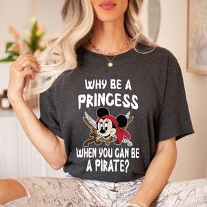K&ouml;nnte beinhalten: Dunkelgraues T-Shirt mit dem Text "WHY BE A PRINCESS WHEN YOU CAN BE A PIRATE?" und einer Cartoon-Piratenmaus. Die Maus tr&auml;gt ein rotes Bandana und h&auml;lt gekreuzte Schwerter.
