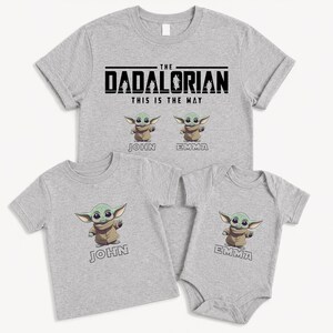 May include: Four light grey t-shirts and a babygrow. The largest shirt reads "THE DADALORIAN THIS IS THE WAY" with Grogu graphics. Smaller shirts and babygrow have Grogu graphics and names. Family matching apparel.