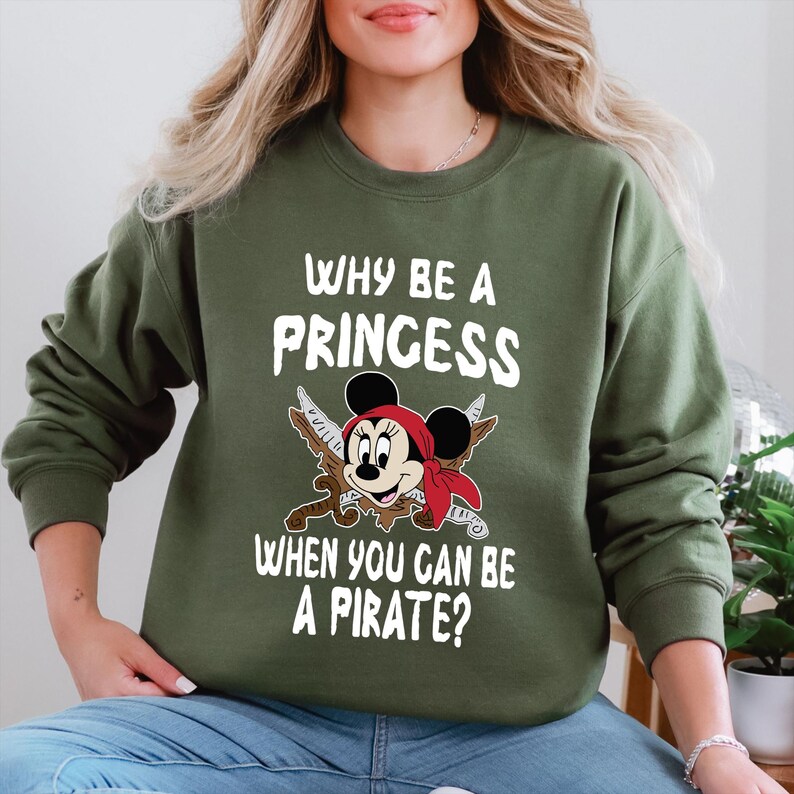 K&ouml;nnte beinhalten: Olivgr&uuml;nes Sweatshirt mit dem Text "WHY BE A PRINCESS WHEN YOU CAN BE A PIRATE?" und einem Cartoon-Bild von Minnie Mouse als Piratin. Minnie Mouse tr&auml;gt ein rotes Bandana und h&auml;lt gekreuzte Schwerter.
